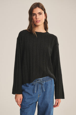 A person stands against a plain background wearing the SAWTELLE CASHMERE SWEATER by Velvet by Jenny Graham, a loose-fitting black ribbed sweater, paired with blue drawstring denim pants.