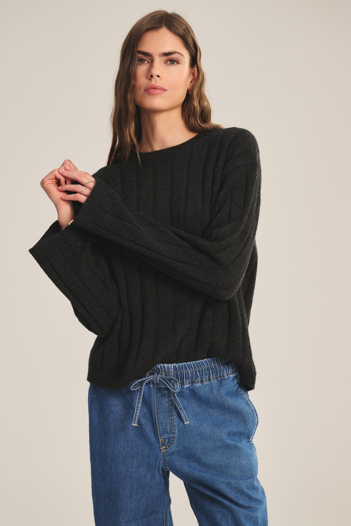 Model wears the SAWTELLE CASHMERE SWEATER by Velvet by Jenny Graham, a relaxed-fit black ribbed knit, paired with blue drawstring jeans against a simple light backdrop.-39838371840193
