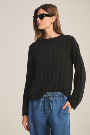 A person wearing black sunglasses, blue drawstring jeans, and the SAWTELLE CASHMERE SWEATER by Velvet by Jenny Graham stands with one hand in their pocket against a plain background.