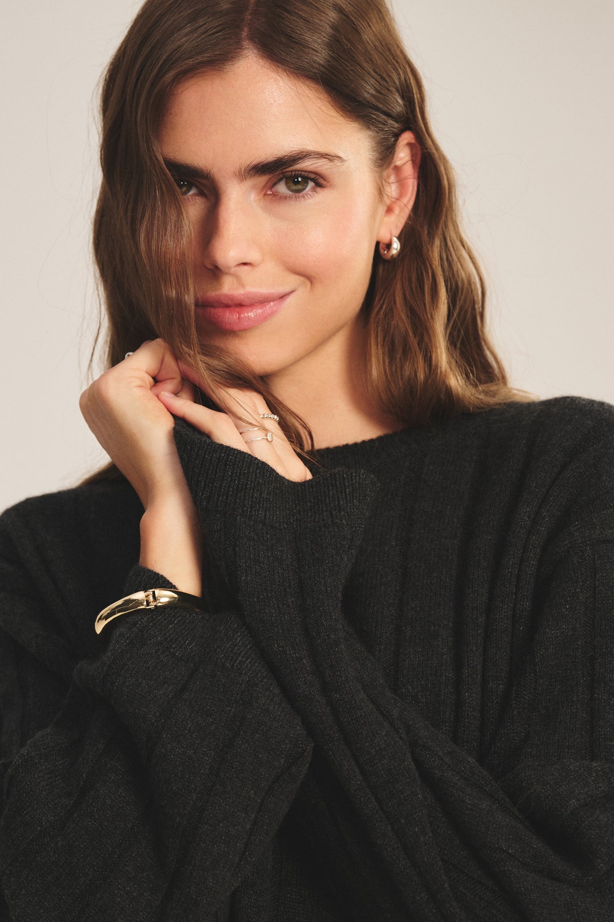 A woman with long brown hair wears the SAWTELLE CASHMERE SWEATER by Velvet by Jenny Graham, paired with gold hoop earrings and a bracelet, standing against a neutral background.-39838371709121