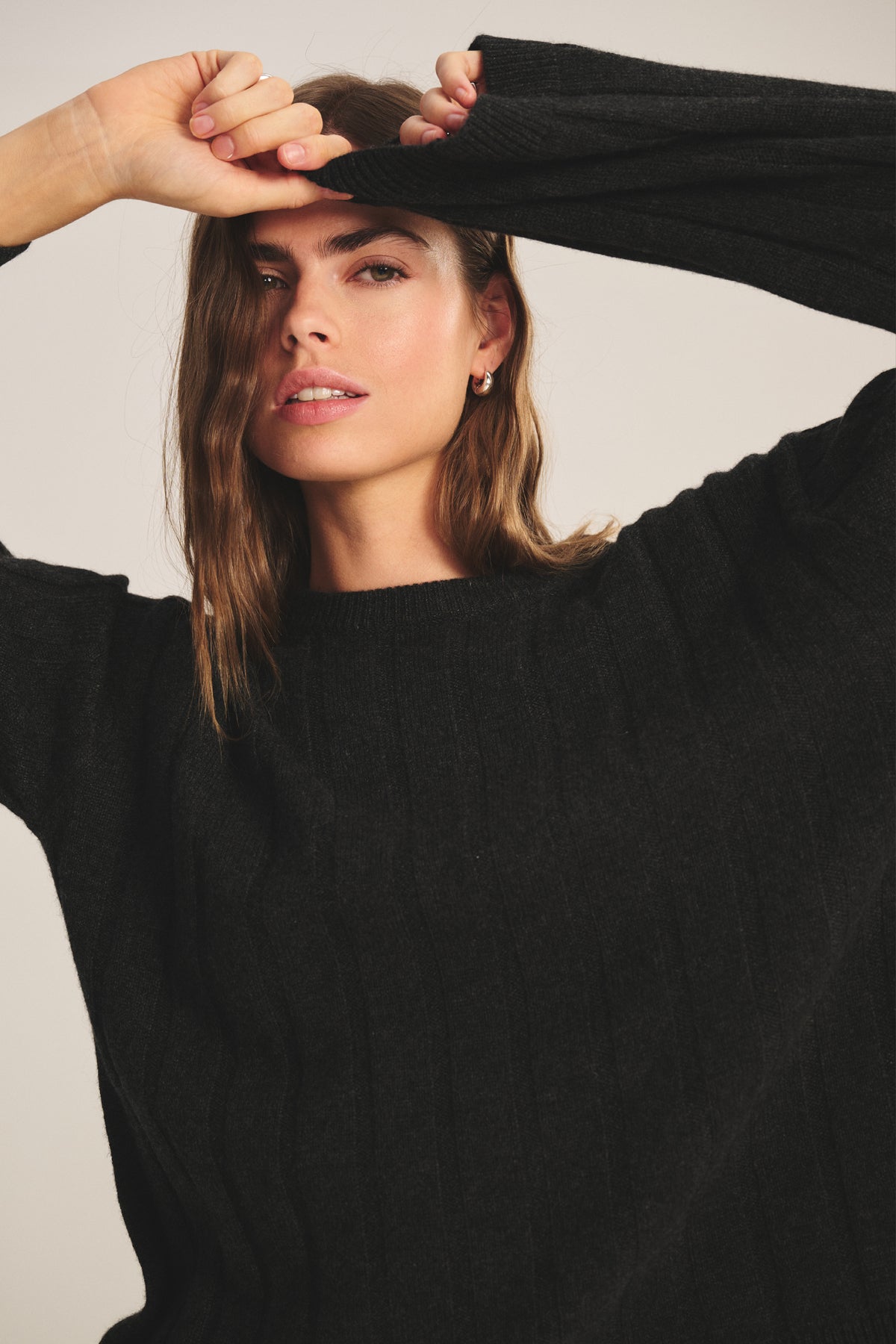   A woman models the SAWTELLE CASHMERE SWEATER by Velvet by Jenny Graham, posing with one arm raised to partially cover her forehead as she looks at the camera. 