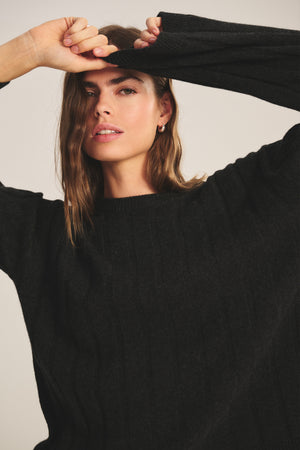 A woman models the SAWTELLE CASHMERE SWEATER by Velvet by Jenny Graham, posing with one arm raised to partially cover her forehead as she looks at the camera.