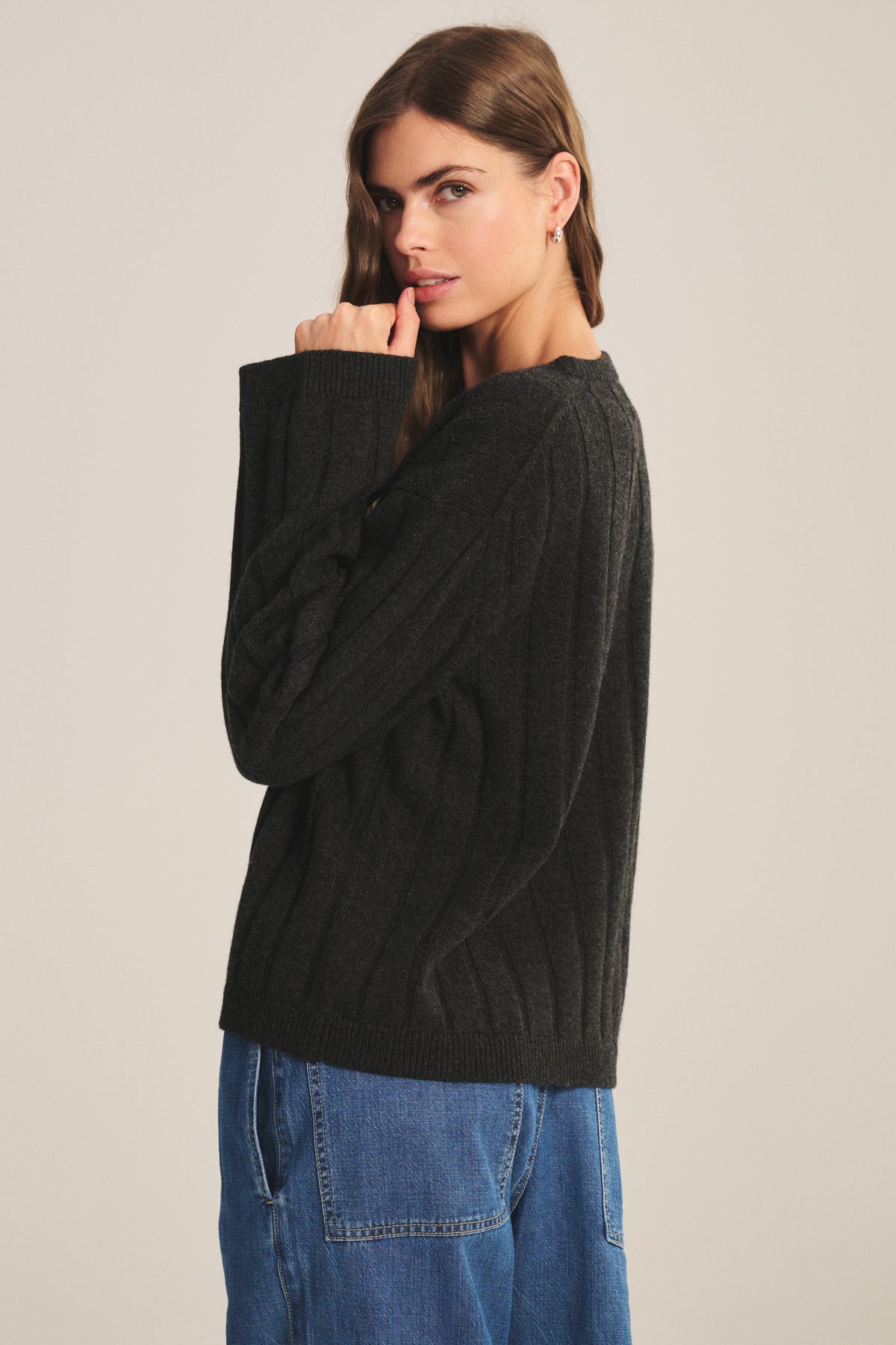 A woman poses sideways against a plain background, wearing the Velvet by Jenny Graham SAWTELLE CASHMERE SWEATER in a dark ribbed knit with blue jeans, gazing at the camera with one hand near her mouth.-39838371610817