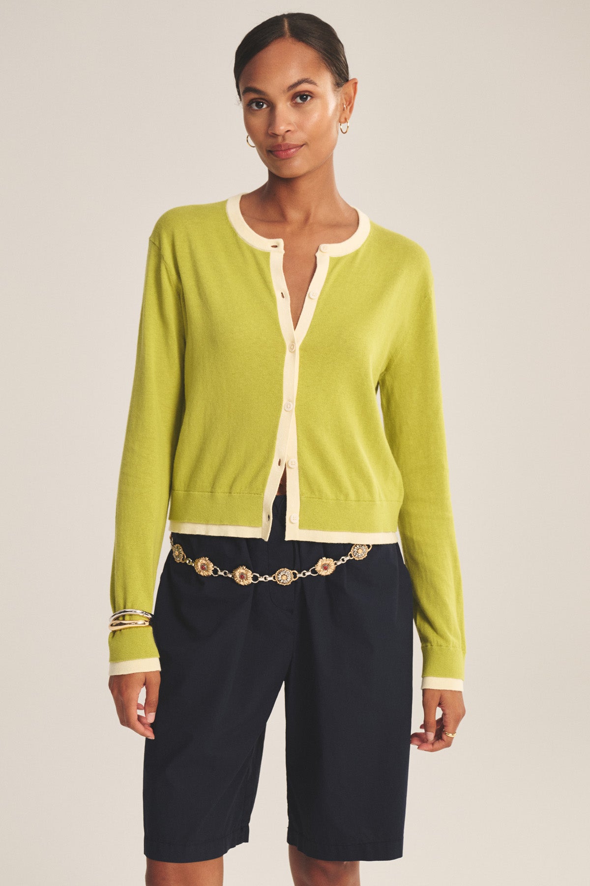 A woman models the SAYLOR CARDIGAN by Velvet by Graham & Spencer in light green with cream trim, paired with navy knee-length shorts, a gold chain belt, hoop earrings, and a bracelet against a plain background.