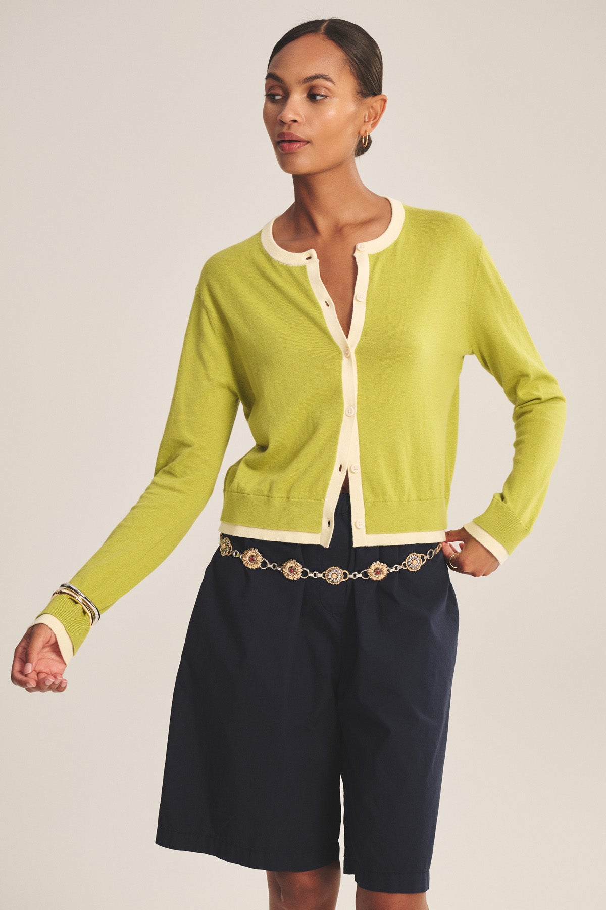 She wears the SAYLOR CARDIGAN by Velvet by Graham & Spencer in light green with cream trim, paired with navy knee-length shorts, a gold chain belt, hoop earrings, and a bracelet against a plain background.-41128865595585