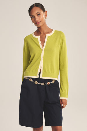 A person wears the Velvet by Graham & Spencer SAYLOR CARDIGAN in light green over a cream top, navy shorts, and a chain belt, standing against a plain background.
