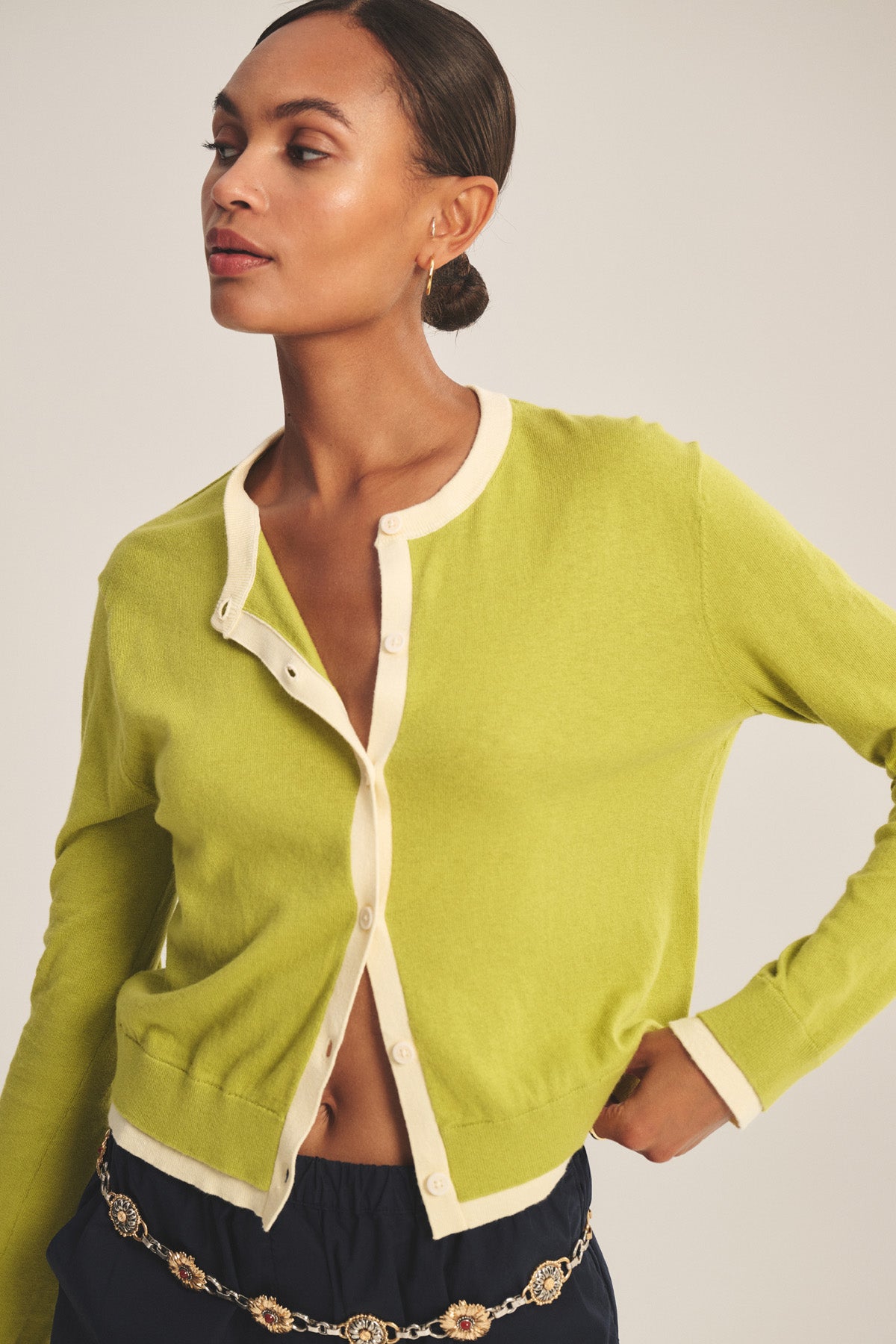 A person wears the SAYLOR CARDIGAN by Velvet by Graham & Spencer—a light green cropped knit with cream trim, unbuttoned at the top and bottom—styled with dark pants and a chain belt against a plain background.-41128865530049