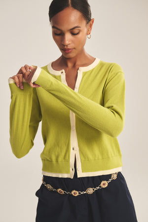 A person models the SAYLOR CARDIGAN by Velvet by Graham & Spencer—a fitted, light green cotton-cashmere piece with cream trim—paired with dark pants and a chain belt, standing against a plain background.