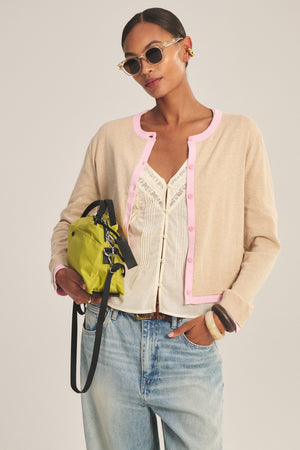 A woman wears the Velvet by Graham & Spencer SAYLOR CARDIGAN with pink trim, a white lace top, light blue jeans, sunglasses, gold earrings, and carries a small green handbag.