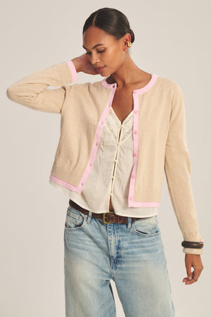 Wearing the SAYLOR CARDIGAN by Velvet by Graham & Spencer—a beige cotton-cashmere piece with pink trim—over a white blouse and blue jeans, the person stands with one hand behind their head, looking down.