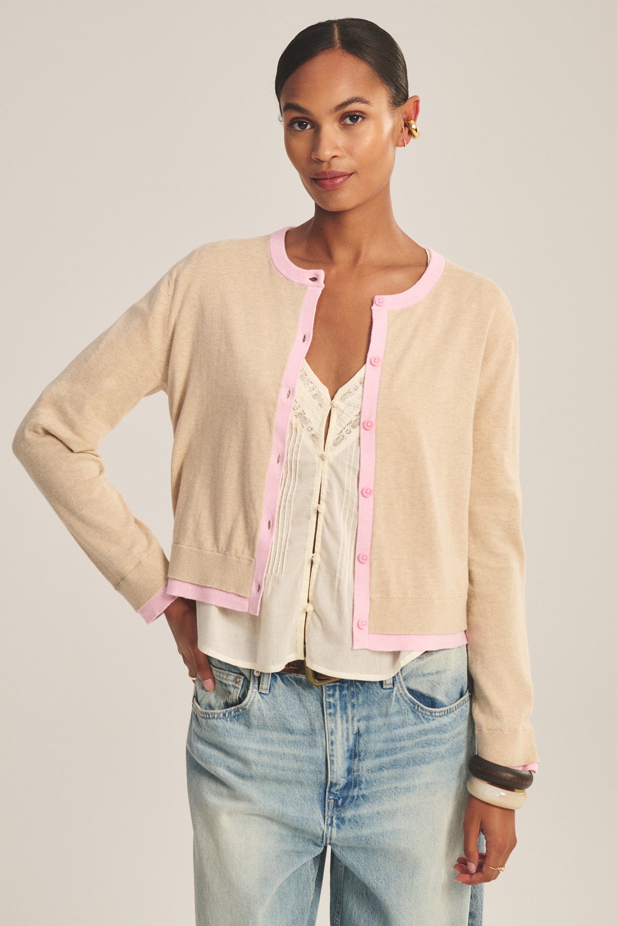A woman stands with one hand on her hip, wearing the SAYLOR CARDIGAN by Velvet by Graham & Spencer—a beige cotton-cashmere cardigan with pink trim—over a white lace top and light blue jeans, against a plain background.-41128865366209
