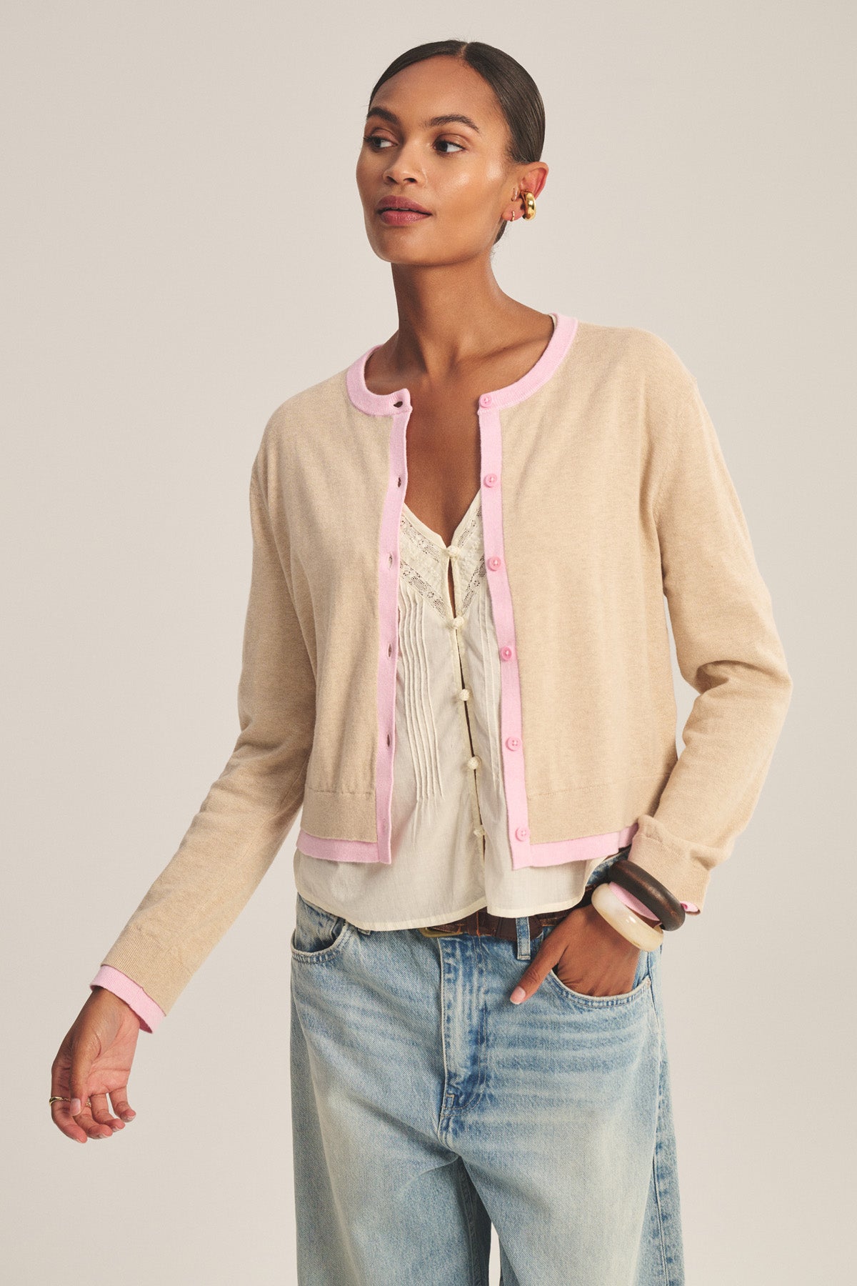 A woman wears the SAYLOR CARDIGAN by Velvet by Graham & Spencer— a cropped cotton-cashmere style with pink trim—over a white top and light blue jeans, standing against a plain background.-41128865333441