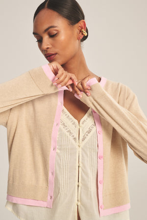 A person wears the SAYLOR CARDIGAN by Velvet by Graham & Spencer—a beige cropped style with pink trim—over a white lace-detailed blouse, standing with eyes closed and arms raised near the face.