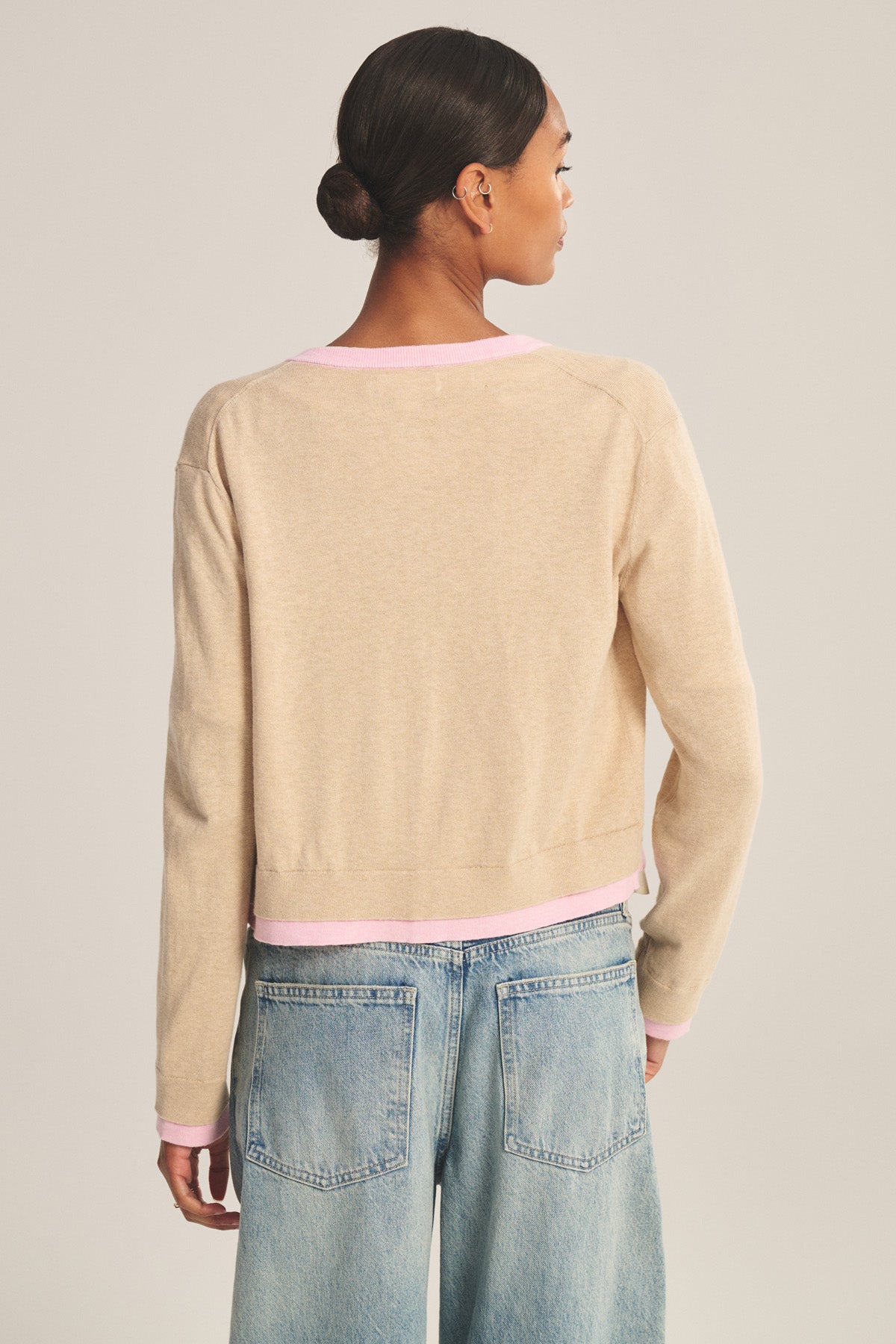 A person with dark hair in a low bun, facing away, is wearing the Velvet by Graham & Spencer SAYLOR CARDIGAN—a beige fitted long-sleeve top with pink trim—paired with light blue jeans.-41128865235137