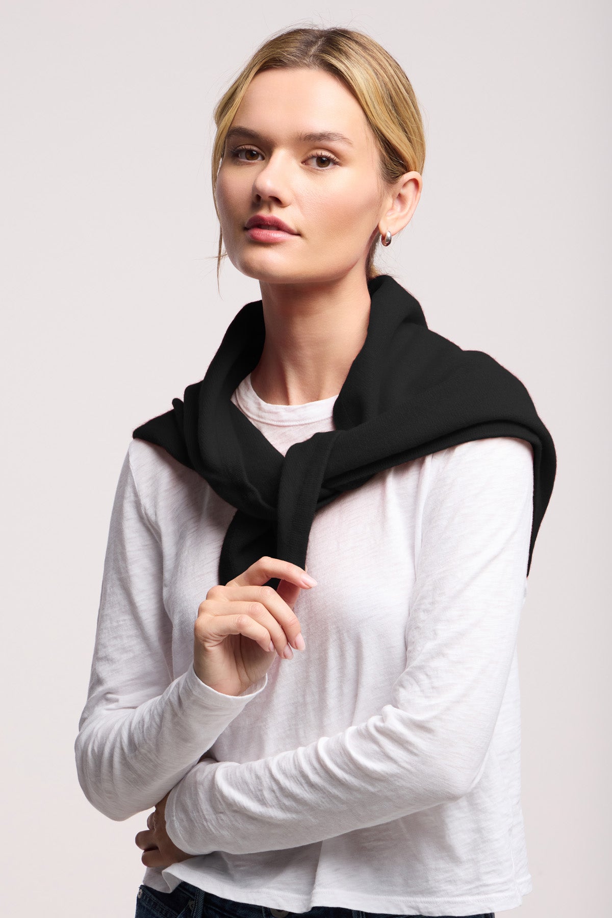 A blonde woman in a white long-sleeve shirt with a black sweater over her shoulders stands against a plain light background, elegantly styled with the Large Triangle Bandana by 40 Colori.-40799605719233