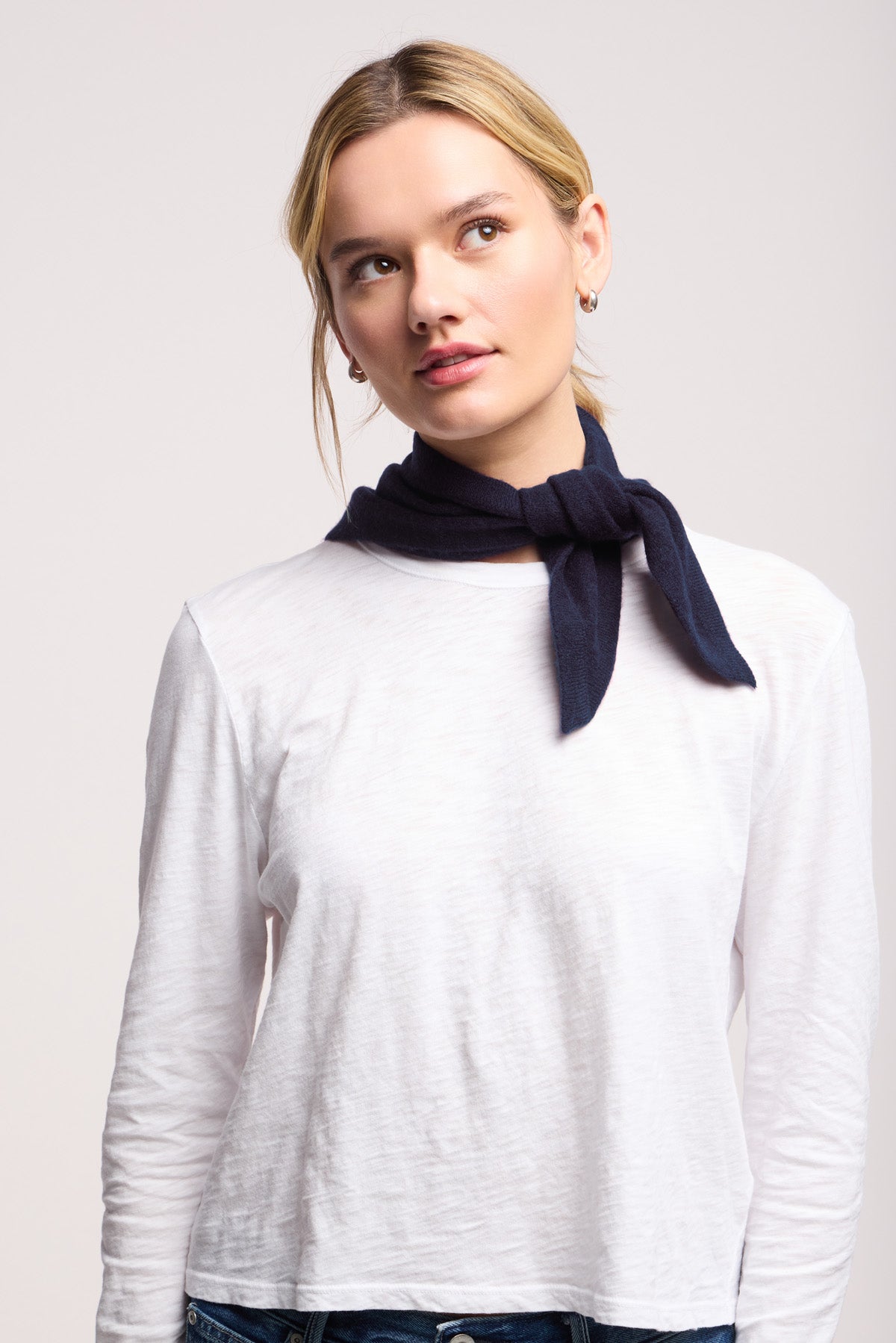 A woman wears the SMALL TRIANGLE BANDANA BY 40 COLORI, a dark scarf from 40 Colori, tied around her neck over a white long-sleeve shirt as she gazes upward against a plain background.-40799579668673