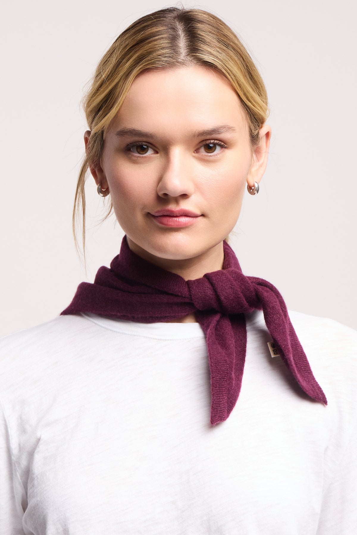 A woman with blonde hair faces the camera against a plain background, wearing a white top and the 40 Colori Small Triangle Bandana in dark burgundy tied around her neck.