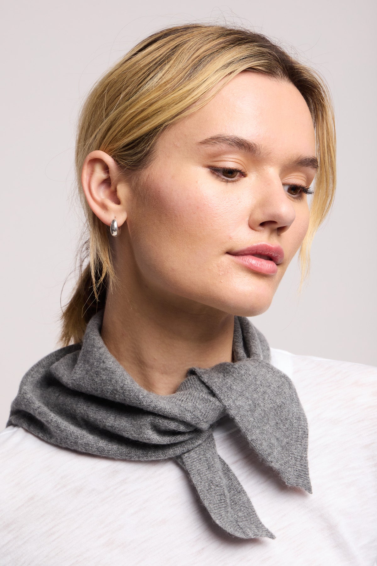 A blonde woman wears the SMALL TRIANGLE BANDANA BY 40 COLORI from 40 Colori around her neck, paired with a white top and small hoop earring, looking slightly downward against a neutral background.-40459073913025