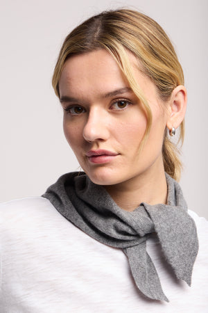 A woman with blonde hair in a low ponytail wears the SMALL TRIANGLE BANDANA BY 40 COLORI, a light grey scarf by 40 Colori, tied around her neck over a white top, looking directly at the camera.