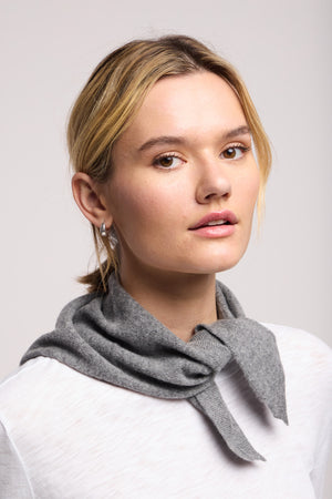 A woman with blonde hair wears the Small Triangle Bandana by 40 Colori in light grey, tied around her neck over a white top, looking at the camera against a plain background.