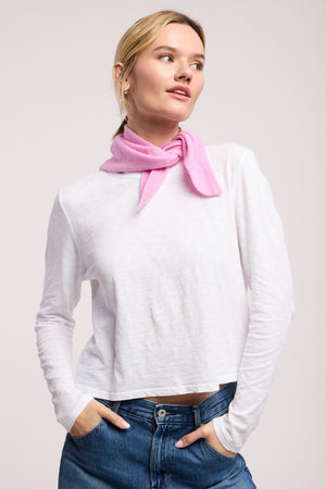 A woman stands against a light background with hands in her pockets, wearing a long-sleeve white shirt, blue jeans, and the SMALL TRIANGLE BANDANA BY 40 COLORI from 40 Colori as a pink scarf around her neck.