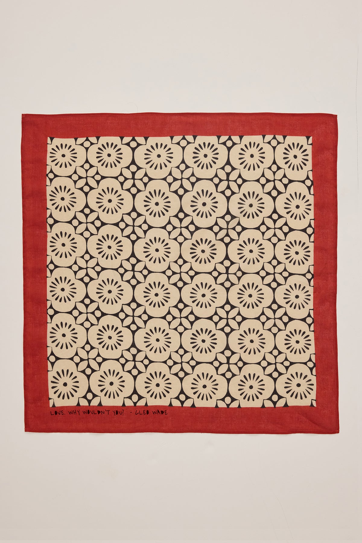 The NOLA SCARF from Velvet by Graham & Spencer showcases a versatile square design featuring a red-bordered bandana with a repeating black floral pattern on a cream backdrop. Made from woven cotton, it bears the inspiring Cleo Wade quote, "Love Will Surmount You," at the bottom edge.-38864648831169