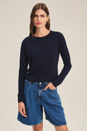 A person models the SCOUT TEE by Velvet by Graham & Spencer, pairing the long-sleeve navy shirt with blue denim shorts and posing with one hand in their pocket against a plain background.