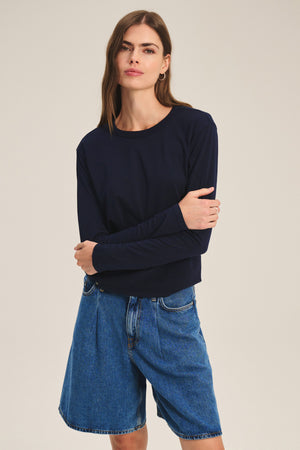 A woman stands against a plain background wearing the SCOUT TEE by Velvet by Graham & Spencer and knee-length blue denim shorts, with her arms loosely crossed.