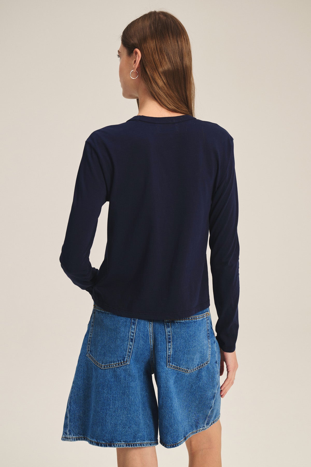 A person with long brown hair, shown from the back against a plain background, wears the SCOUT TEE by Velvet by Graham & Spencer—a navy long-sleeve shirt paired with denim shorts.
