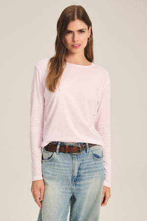 A woman wears the SCOUT TEE by Velvet by Graham & Spencer in light pink, paired with blue jeans and a brown belt, standing facing forward against a plain background.