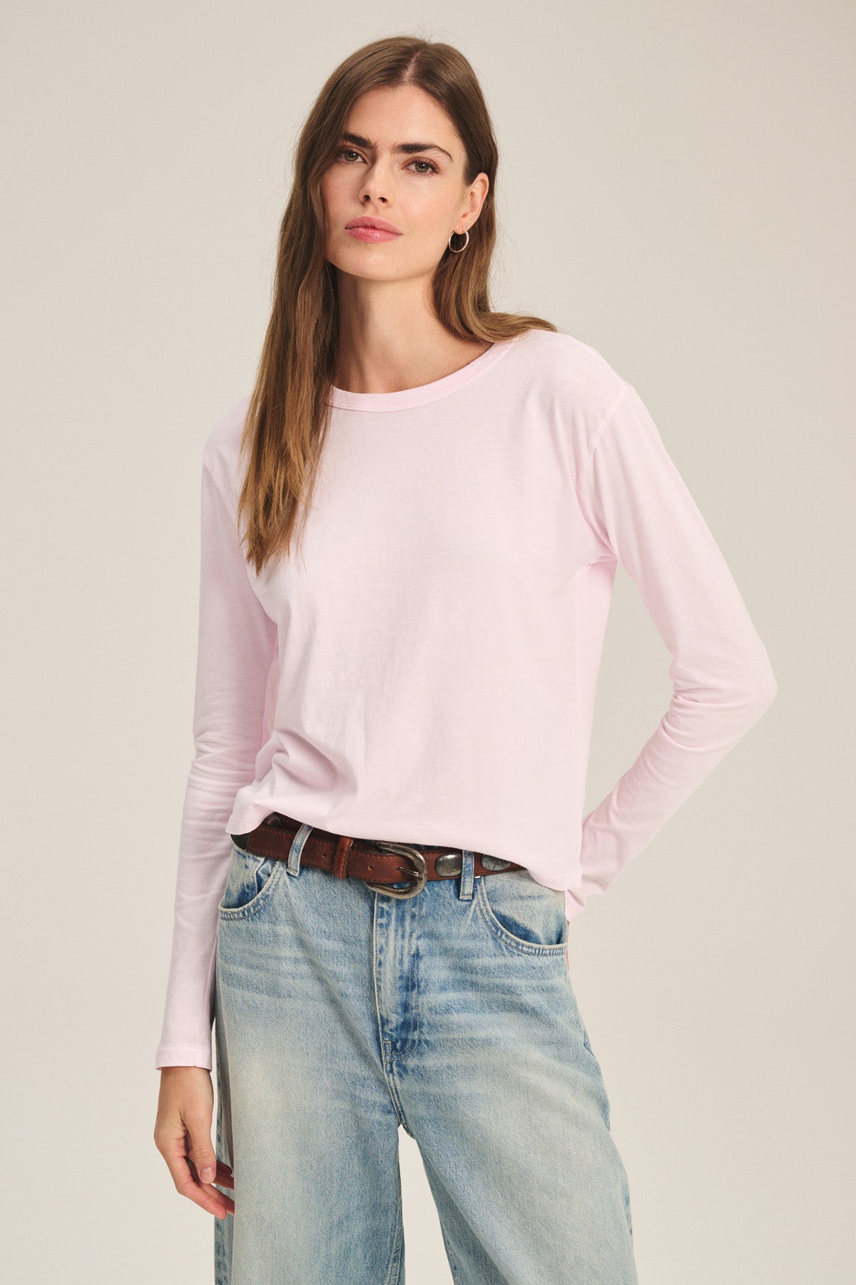 A woman wears the SCOUT TEE by Velvet by Graham & Spencer—a light pink long-sleeve shirt—tucked into light blue jeans with a brown belt, standing against a plain background.