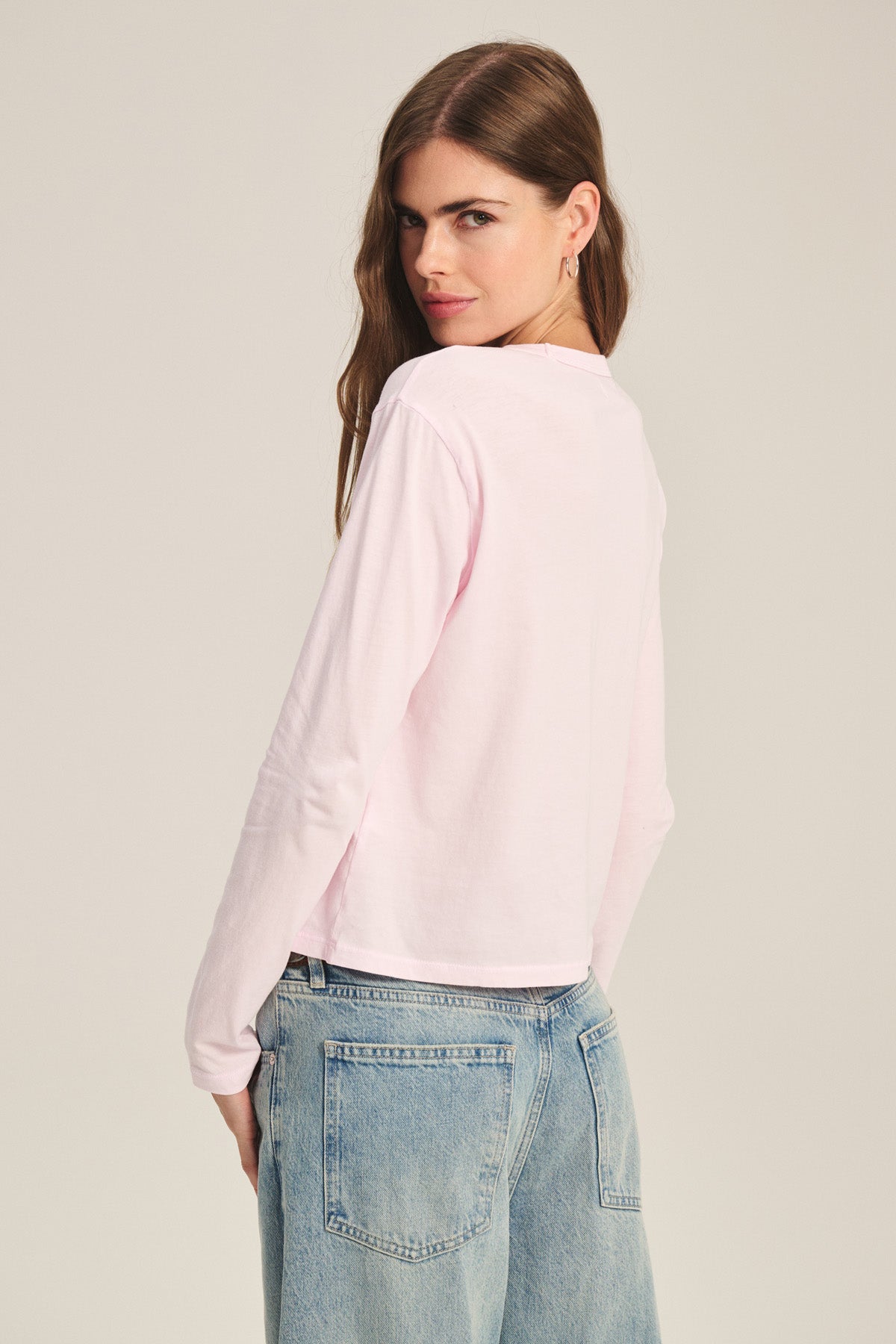 A woman wearing the Velvet by Graham & Spencer SCOUT TEE in light pink and light blue jeans stands with her back to the camera, looking over her shoulder.