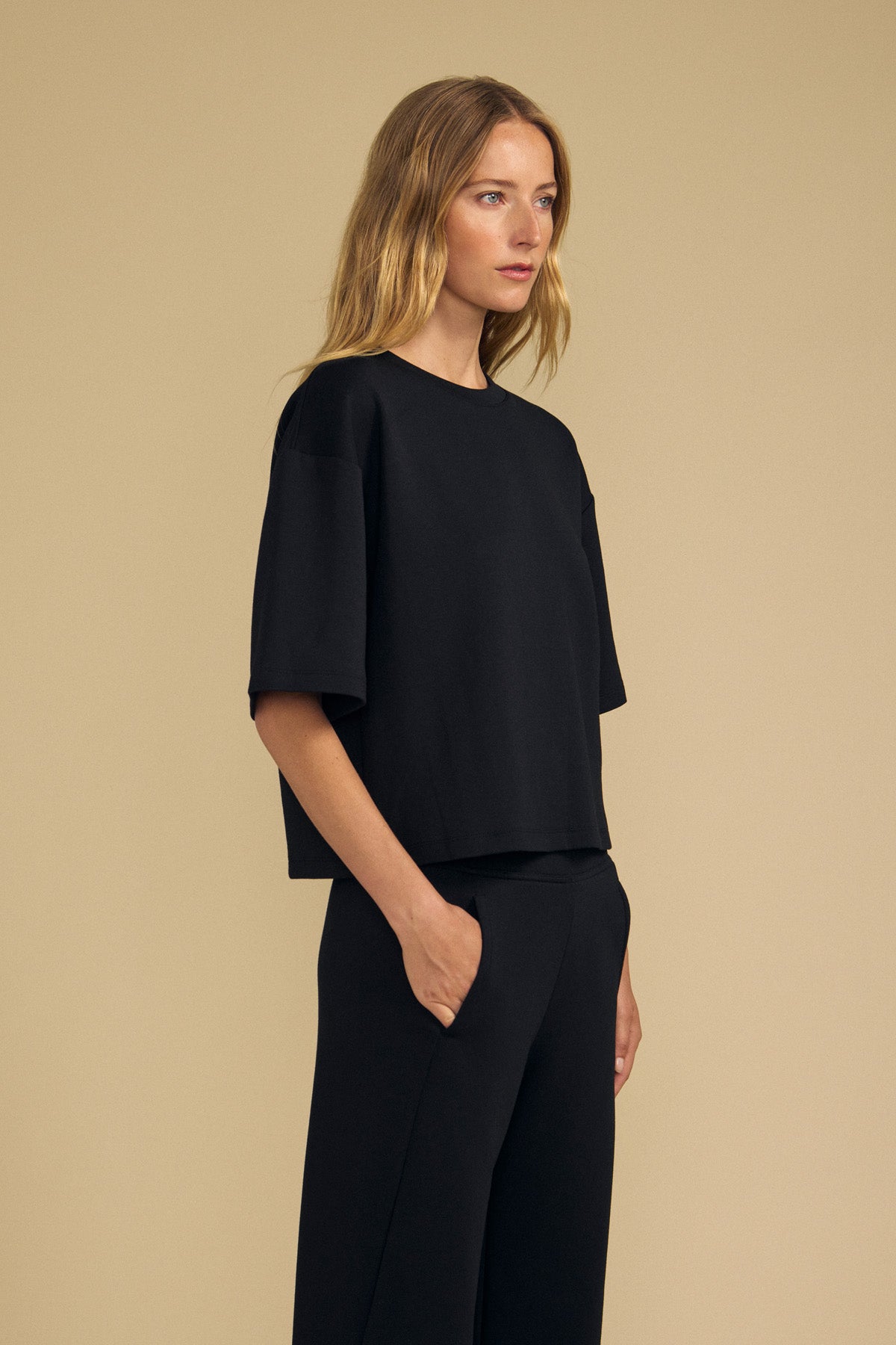 A woman stands against a beige background wearing the ALIZZA TOP by Velvet by Graham & Spencer—a loose-fitting black T-shirt with short sleeves—paired with matching black pants, her right hand in her pocket.