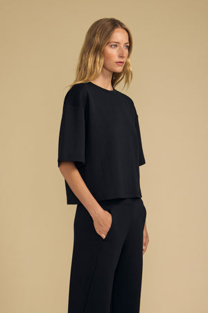 A woman stands against a beige background wearing the ALIZZA TOP by Velvet by Graham & Spencer—a loose-fitting black T-shirt with short sleeves—paired with matching black pants, her right hand in her pocket.