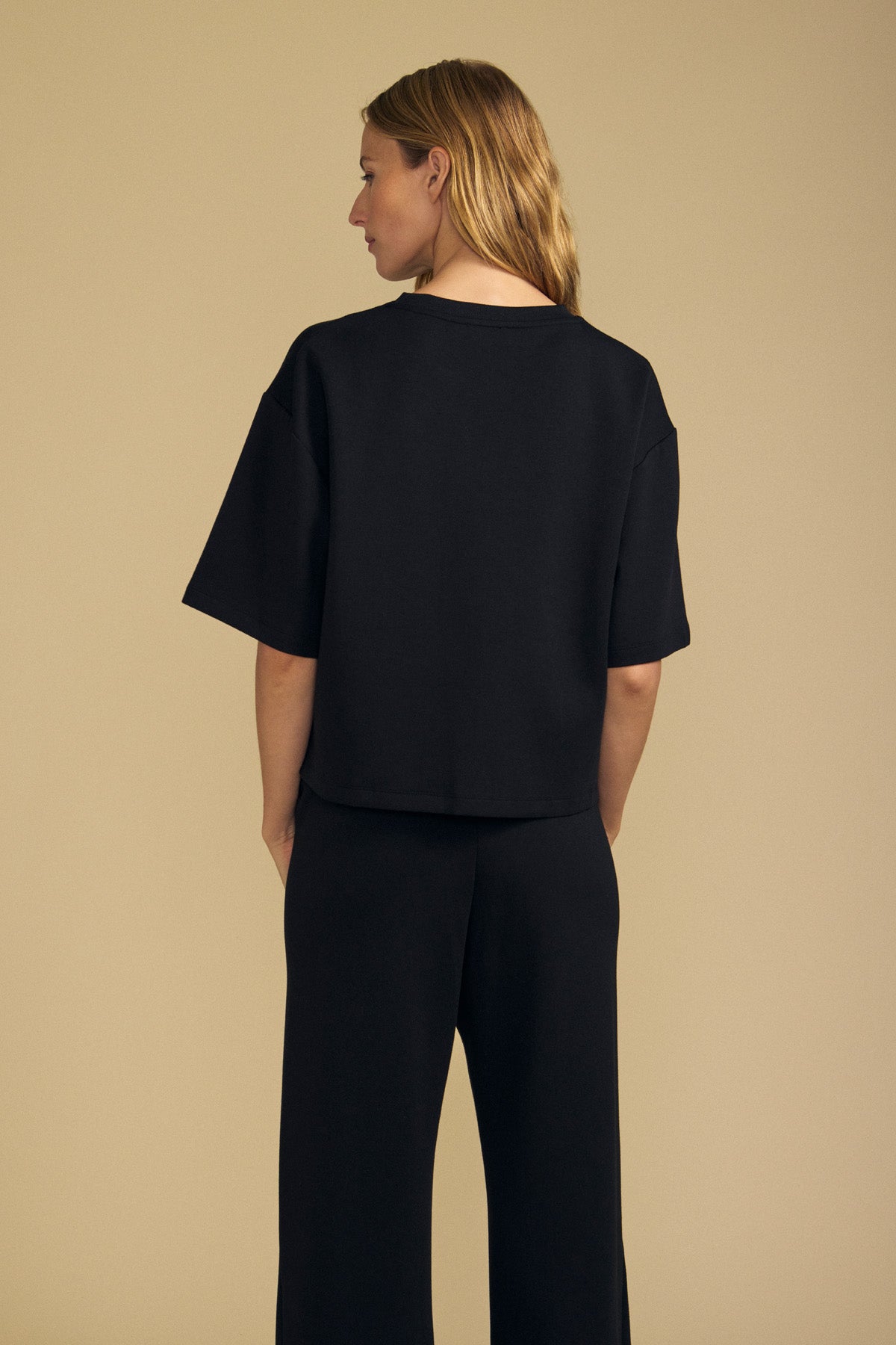 A person with shoulder-length blond hair wears the Velvet by Graham & Spencer ALIZZA TOP in black, paired with matching black pants, facing away against a plain beige background.