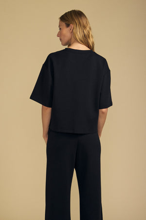 A person with shoulder-length blond hair wears the Velvet by Graham & Spencer ALIZZA TOP in black, paired with matching black pants, facing away against a plain beige background.