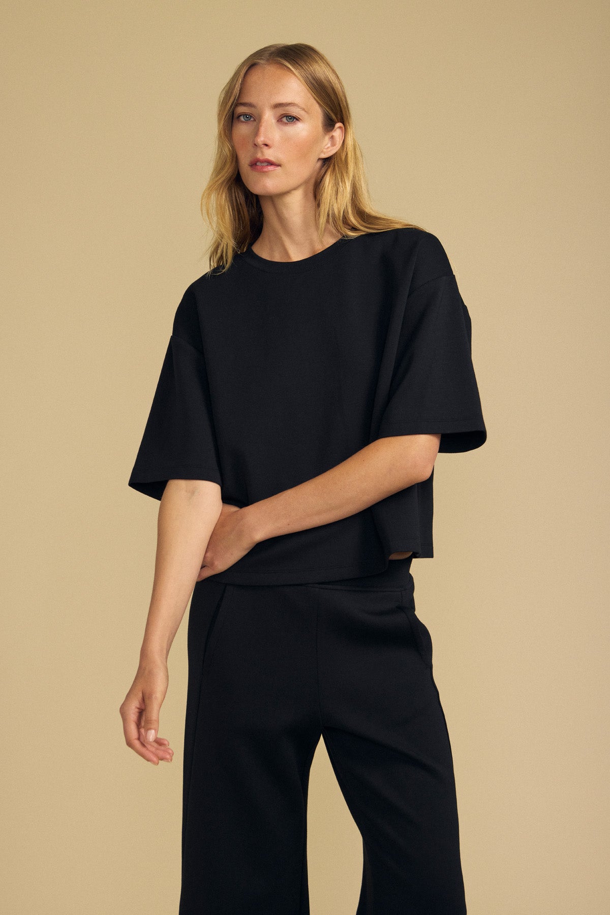 A person with long blonde hair wears the Velvet by Graham & Spencer ALIZZA TOP, a loose-fitting black short-sleeve top, paired with black pants and stands against a plain beige background.-41813566554305