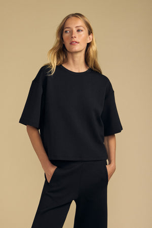 A person stands against a beige background, wearing the loose-fitting black ALIZZA TOP by Velvet by Graham & Spencer with matching black pants, one hand in their pocket.