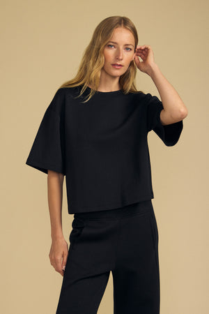 A woman with long blonde hair wears the loose-fitting black ALIZZA TOP by Velvet by Graham & Spencer, along with matching black pants, standing against a beige background.