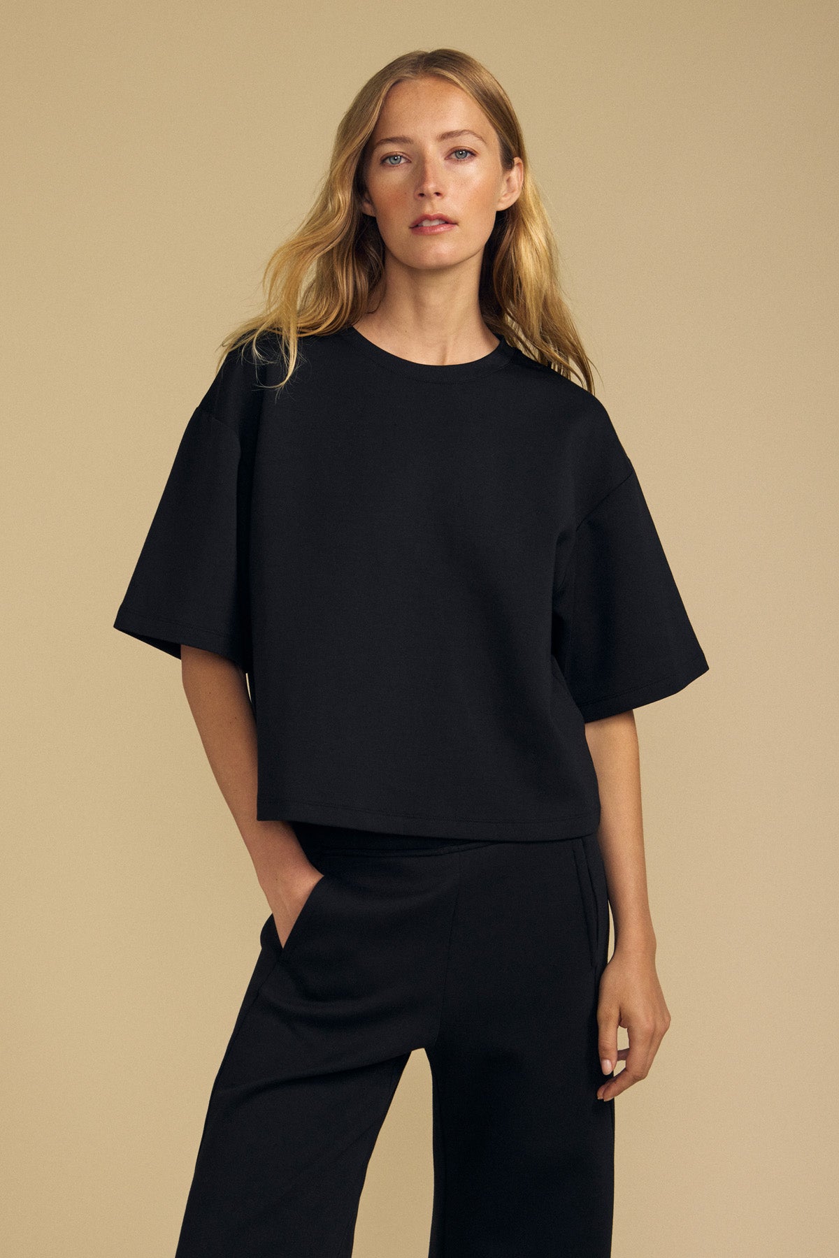 A woman with long blonde hair wears the loose-fitting ALIZZA TOP by Velvet by Graham & Spencer in black, paired with matching black pants, standing against a plain beige background.