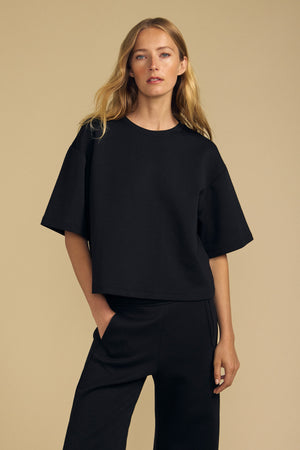 A woman with long blonde hair wears the loose-fitting ALIZZA TOP by Velvet by Graham & Spencer in black, paired with matching black pants, standing against a plain beige background.