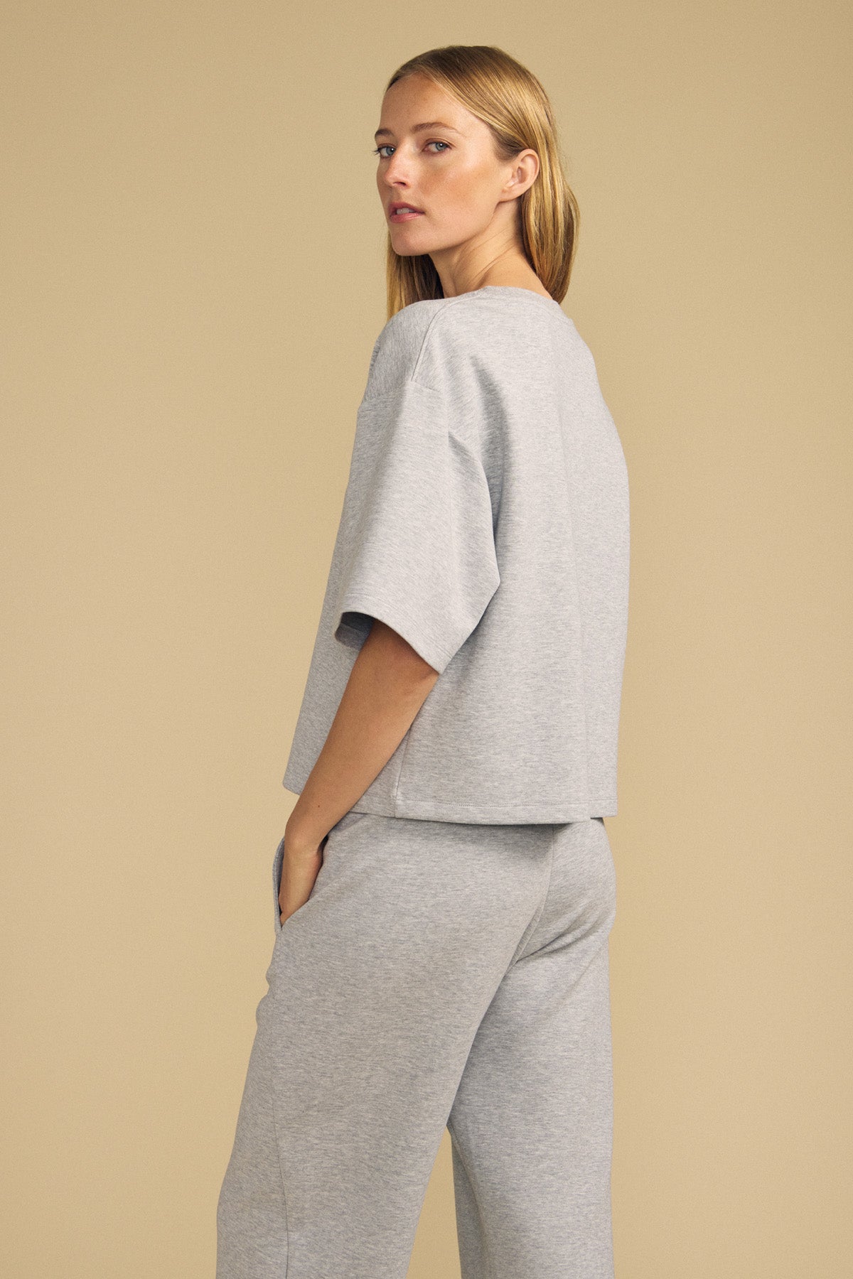 A woman wearing the Velvet by Graham & Spencer ALIZZA TOP in light gray, paired with matching pants, stands sideways against a beige background.