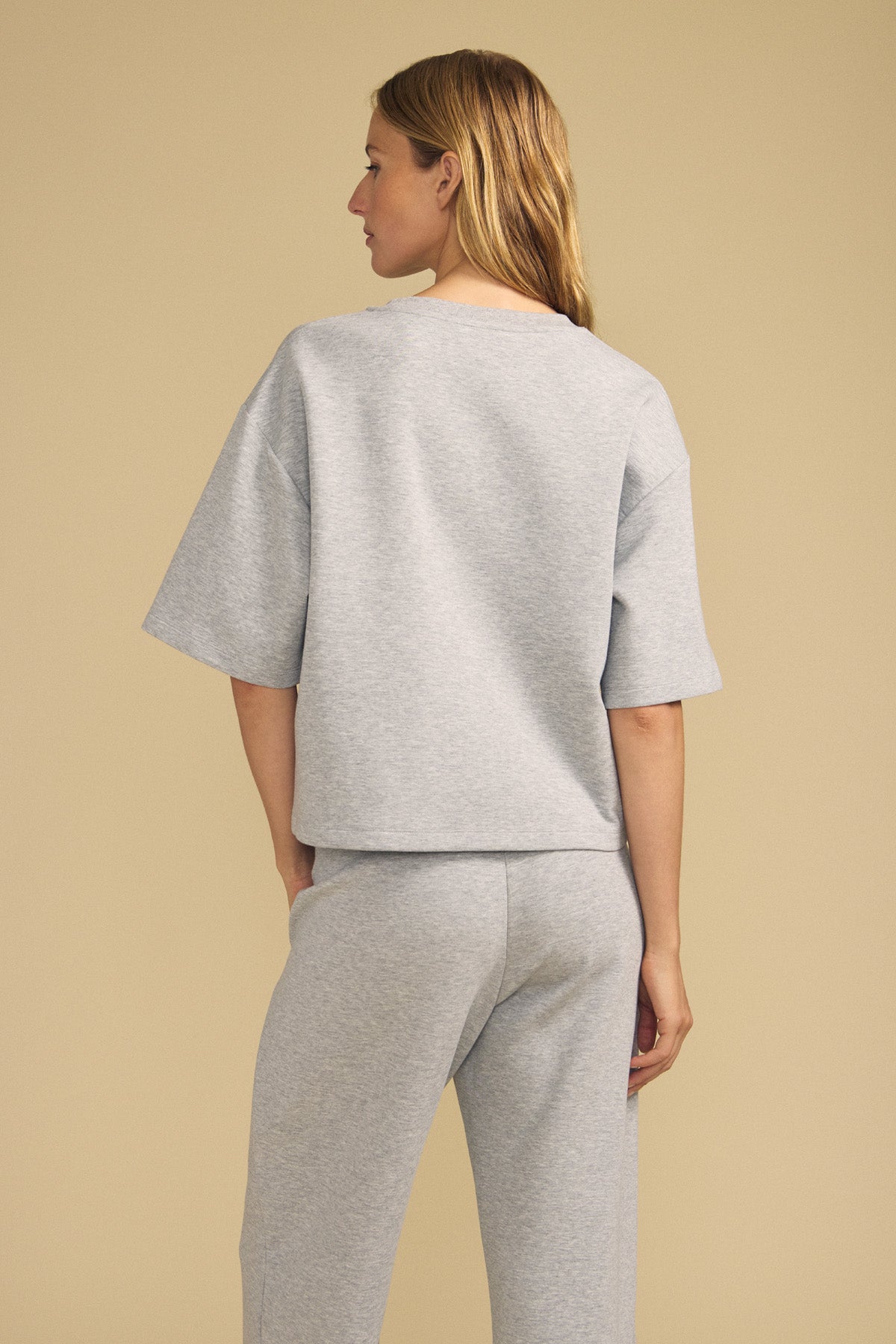 A person stands facing away against a beige background wearing the Velvet by Graham & Spencer ALIZZA TOP, a loose-fitting, light gray short-sleeve top paired with matching pants.