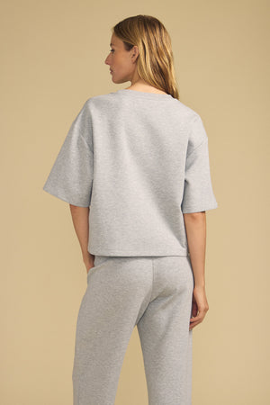 A person stands facing away against a beige background wearing the Velvet by Graham & Spencer ALIZZA TOP, a loose-fitting, light gray short-sleeve top paired with matching pants.