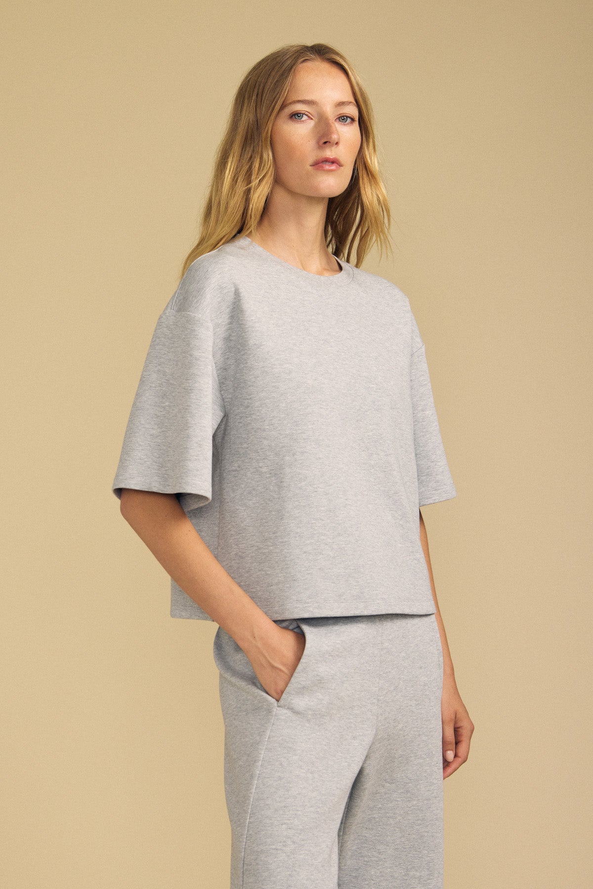 A woman with long blonde hair wears the ALIZZA TOP by Velvet by Graham & Spencer—a loose-fitting, light gray short-sleeve top paired with matching pants—while standing against a neutral beige background.-41813566324929