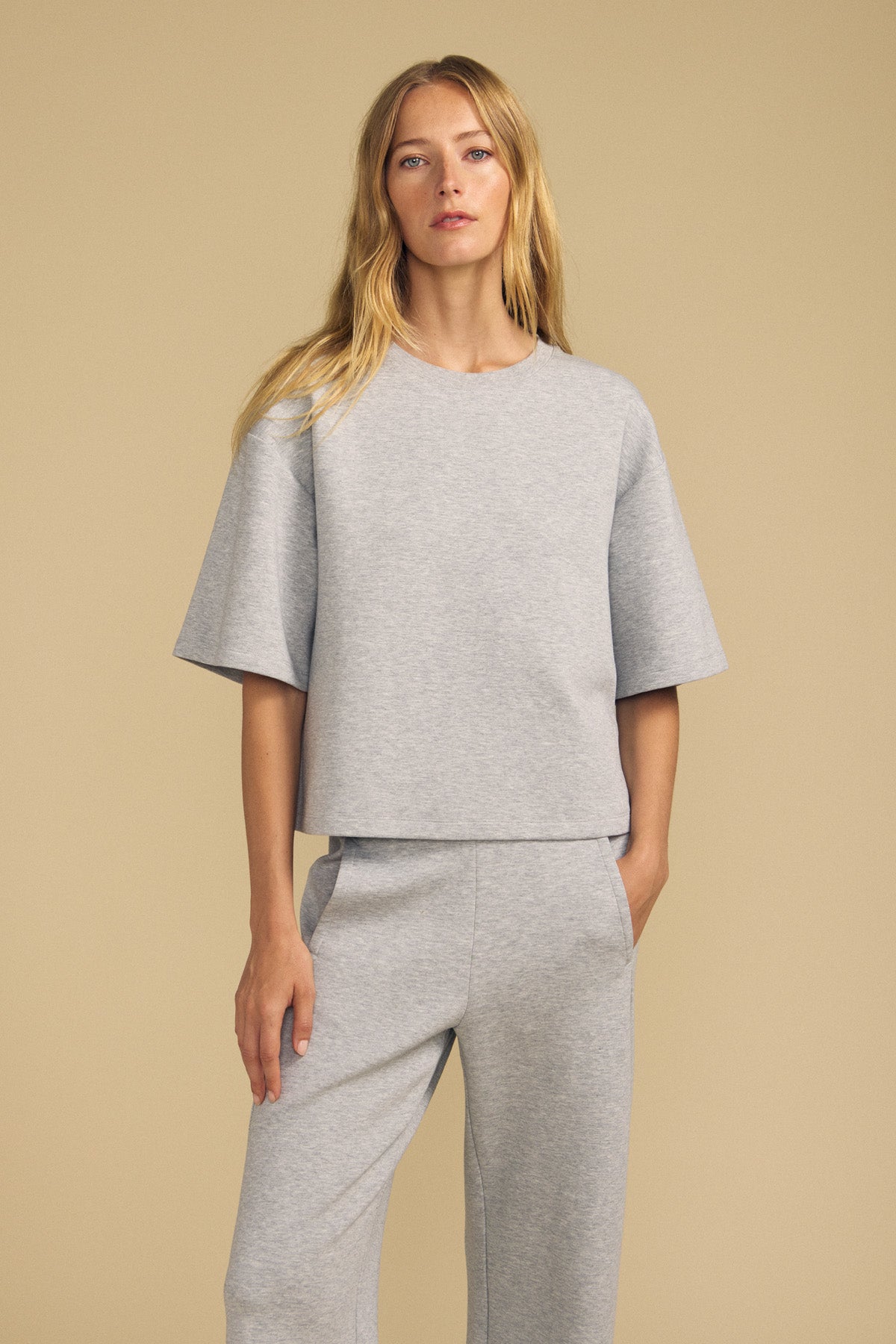 A woman stands against a beige background wearing the ALIZZA TOP by Velvet by Graham & Spencer, a loose-fitting, light gray short-sleeve top paired with matching pants, with her hand in her pocket.