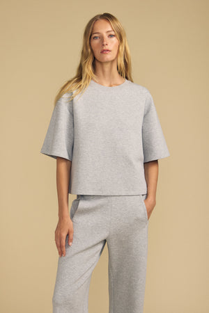 A woman stands against a beige background wearing the ALIZZA TOP by Velvet by Graham & Spencer, a loose-fitting, light gray short-sleeve top paired with matching pants, with her hand in her pocket.