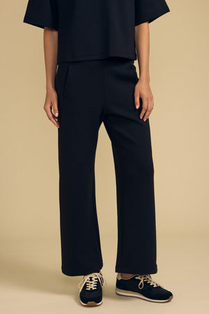 Wearing the Velvet by Graham & Spencer ARA SWEATPANT in black, paired with a matching black top and black sneakers with white soles, the person stands against a plain beige background, with only their lower half visible.