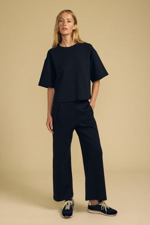 A woman stands against a beige backdrop, wearing the ARA SWEATPANT by Velvet by Graham & Spencer—a pair of loose black wide-leg pants—paired with a matching short-sleeve top and black sneakers with white laces. She looks forward with a neutral expression.