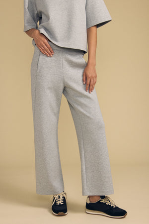 A person is wearing the Velvet by Graham & Spencer ARA SWEATPANT—a pair of loose, wide-leg gray pants—paired with a matching loose gray short-sleeve top and black sneakers with beige laces, standing against a neutral beige background.